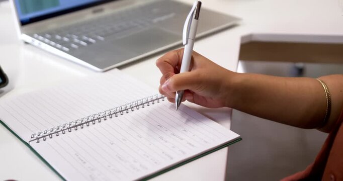 Adult woman taking notes using laptop at white desk, writing with white pen in spiral notebook
