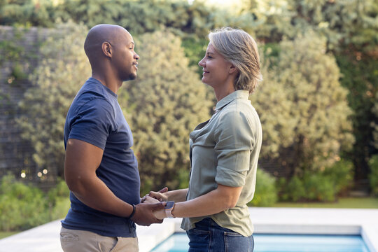 Diverse couple holding hands at backyard pool, senior woman with smartwatch, man with wristband
