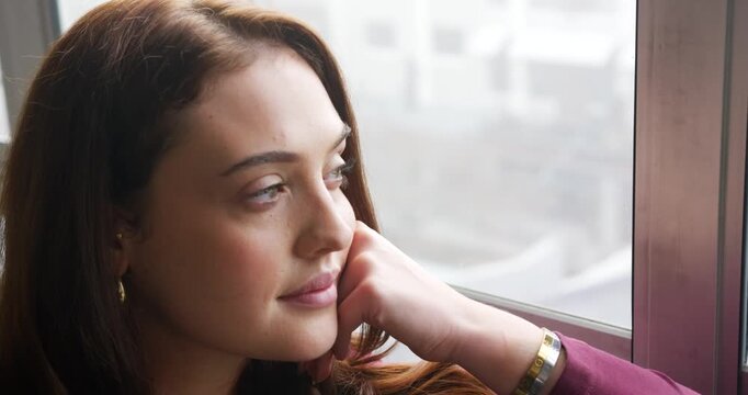 Woman resting chin on right hand, gazing out big window, daylight drawing gaze, gold bangle