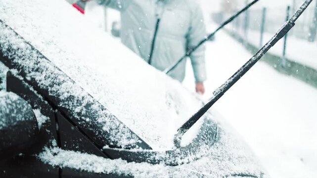 Close-up of a person clearing fresh white snow off an automobile front window and wipers after blizzard. Winter vehicle maintenance, automotive care, and freezing weather concept.