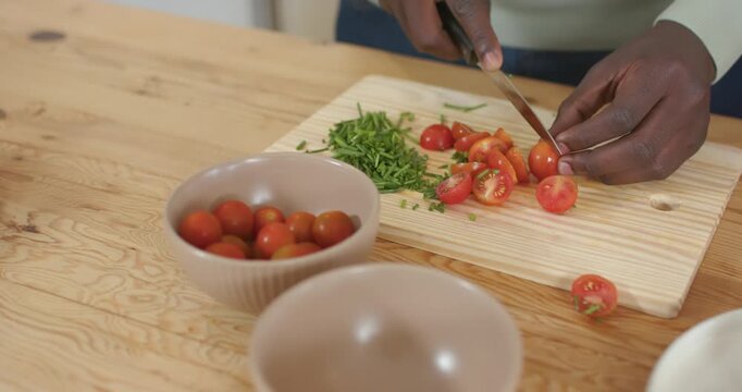 African American woman placing cherry tomato on cutting board, slicing with knife for salad prep