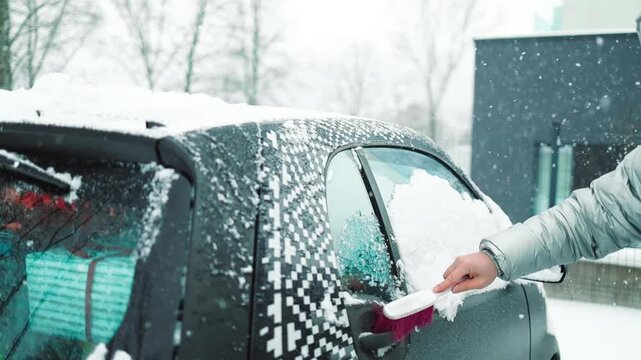 Close-up of a person clearing fresh white snow off an automobile front window and wipers after blizzard. Winter vehicle maintenance, automotive care, and freezing weather concept.