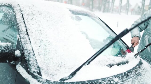 Close-up of a person clearing fresh white snow off an automobile front window and wipers after blizzard. Winter vehicle maintenance, automotive care, and freezing weather concept.