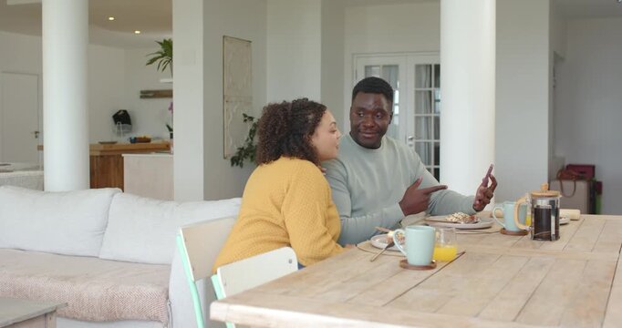 African American couple sharing smartphone, sitting at wooden dining table, leaning and hugging
