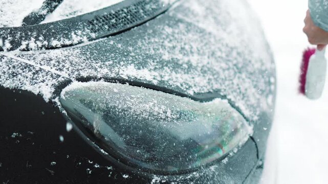 Close-up of a person clearing fresh white snow off an automobile front window and wipers after blizzard. Winter vehicle maintenance, automotive care, and freezing weather concept.