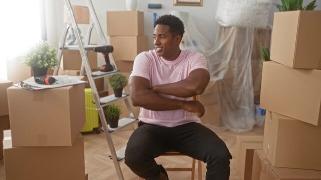 Man sitting smiling with crossed arms, wearing pink t shirt, surrounded by stacked cardboard boxes, ladder and power drill in building; pride fresh start.