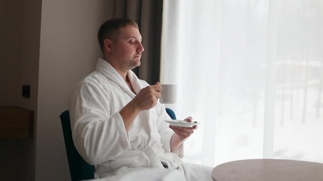 Side view profile of a relaxed millennial male holding a cup of tea while sitting in an armchair near a large window. Concept of morning routine, relaxation, luxury spa wellness, and hospitality