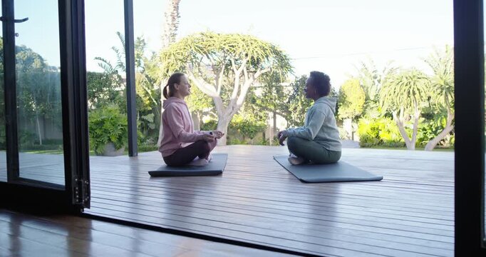 Diverse women in hoodies meditating on mats on deck by glass doors, breathing then stretching up