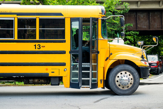 Yellow school bus with its passenger door open pauses beneath a railroad bridge on a suburban street, illustrating daily student transportation, neighborhood bus routes, and public education services.