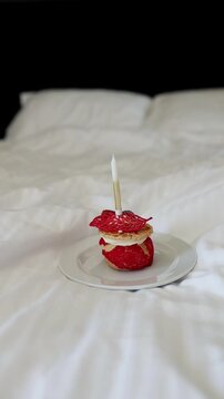 Small delicious strawberry cake or pastry with a lit candle standing on a plate on white sheets in bedroom. Concept of birthday celebration, anniversary surprise, and romantic morning in hotel.