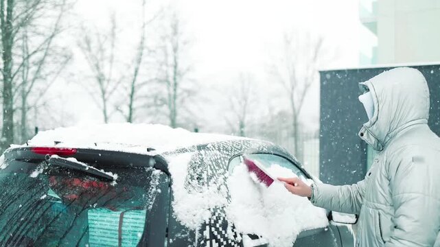 Close-up of a person clearing fresh white snow off an automobile front window and wipers after blizzard. Winter vehicle maintenance, automotive care, and freezing weather concept.