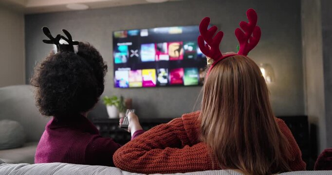 Female pals wearing antlers on couch using remote navigating TV menu choosing show sharing popcorn