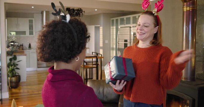 Diverse female friends exchanging wrapped gift for holiday, hugging and checking bow in kitchen