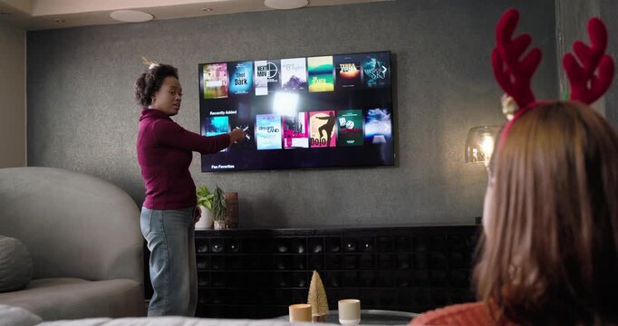 Female friends in living room, standing woman tapping TV to select movie, dancing, donning antlers