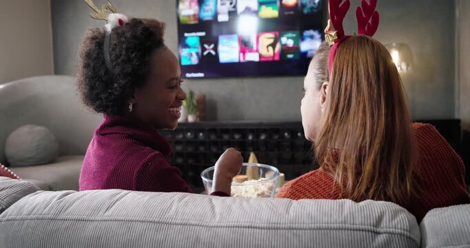 Diverse female friends wearing antlers watching TV grid to choose show, sharing popcorn on couch