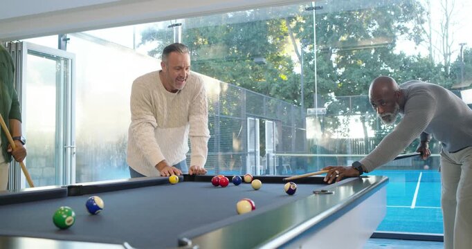 Diverse men at home watching center man leaning on pool table, shooter striking cueball playing