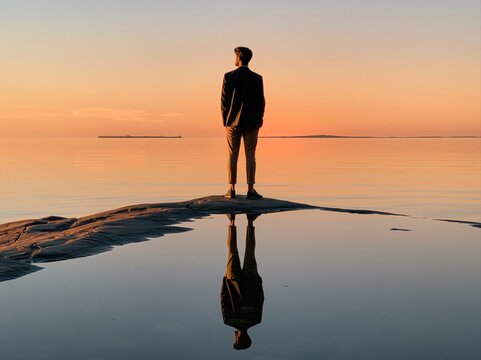 Person stands on rock by water looking at sunset over calm sea in quiet evening light