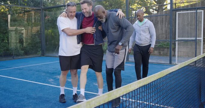 Diverse male teammates steadying limping teammate while walking to gate on padel court with paddles