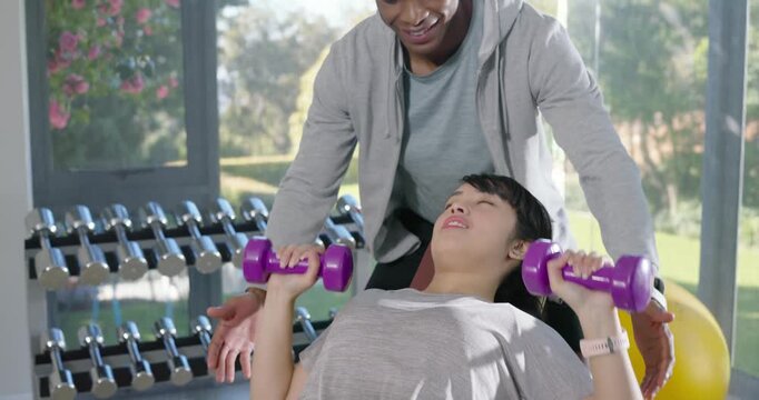 Male trainer positioning, spotting woman pressing weights on incline bench at gym ensuring form