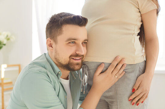 Happy couple expecting baby. Young married man and woman parents to be awaiting baby. Young father puts his ear to belly of his pregnant wife to feel baby kicking. Pregnancy, love, family concept