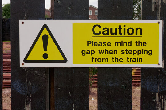 Railway sign, Caution. Please mind the gap when stepping from the train. Modern in bright white, yellow and black with bold, graphic design Attached to a wooden fence with a rail way shunting yard beh