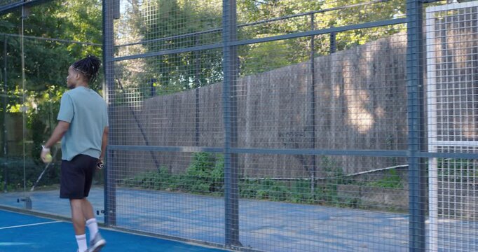 Couple entering blue paddle court, woman exiting gate, African male warming up with paddle and ball