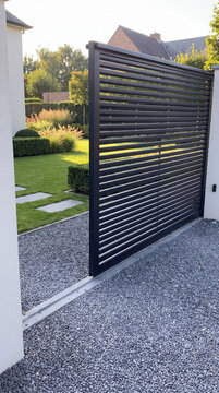 Modern black metal sliding gate with horizontal slats. Landscaped garden with green lawn and gravel driveway. Vertical photo