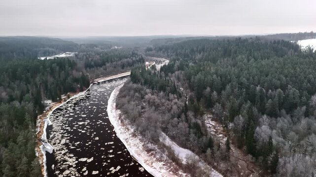 Aerial view of a winding river filled with ice floes flowing through a dense, snow-covered forest landscape in Latvia.
