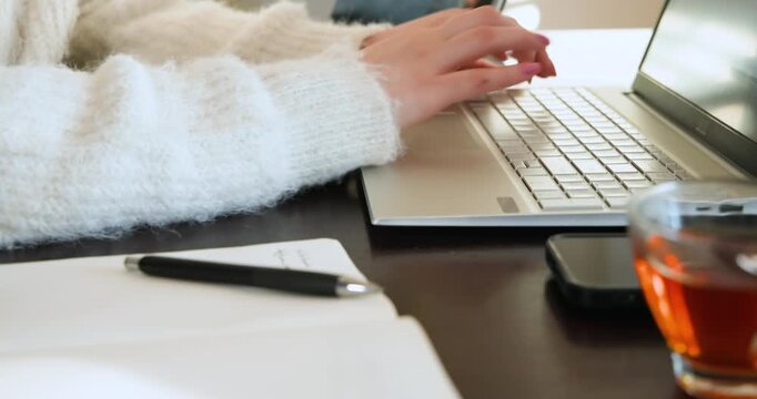 Female hands in fluffy sweater typing on laptop pausing reaching phone writing notes at desk