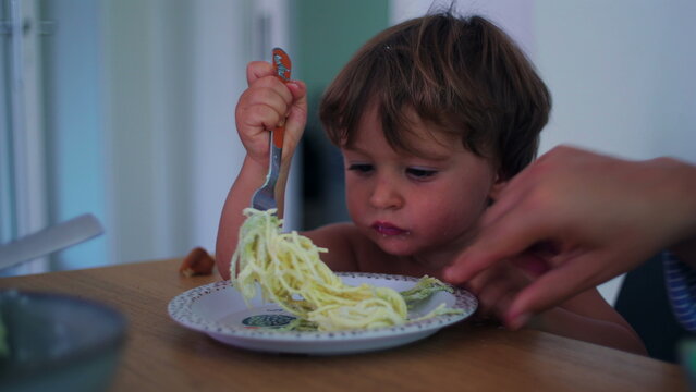 Child twirling spaghetti with fork at the kitchen table at home, focused self-feeding practice representing motor development, confidence and healthy independence