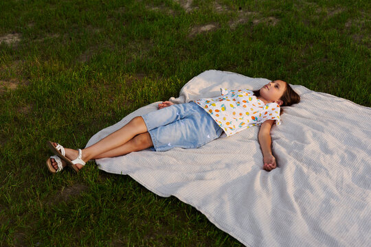 A child lies on a blanket in a grassy area, relaxing and watching the sky. The scene shows simple enjoyment on a clear day with sunshine