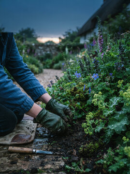 Person in denim overalls kneels in a garden bed working soil near a stone building
