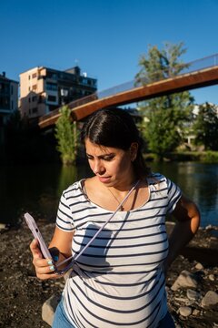 Woman uses phone outdoors by river with bridge and buildings