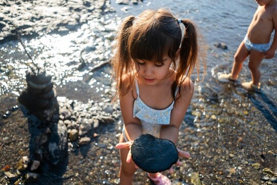 A young white girl holds a black rock by the water's edge