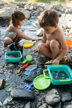 A white boy and girl play with water and toys by a rocky stream