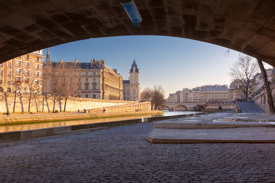 Riverside view in central Paris framed by a stone bridge, with historic buildings, quays and another bridge along the Seine River.
