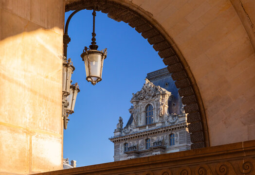Ornate historic facade with sculptural details and hanging lanterns seen through a large stone arch in Paris, France.