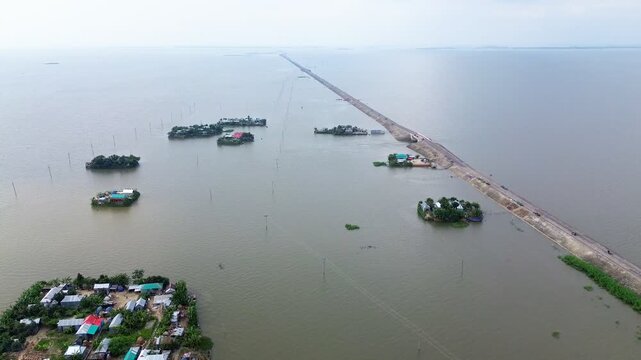 Aerial view of a flooded landscape with a long embankment road and small clusters of houses on elevated land in Kishoreganj, Dhaka Division, Bangladesh.