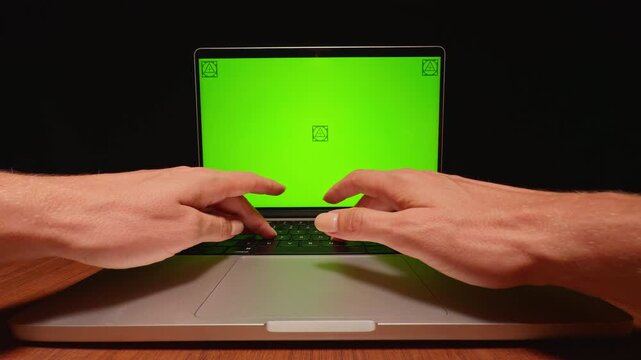 Hands Type on a Laptop Keyboard Before a Green Screen Display on a Wooden Desk, Supporting Technology and Remote Work Content