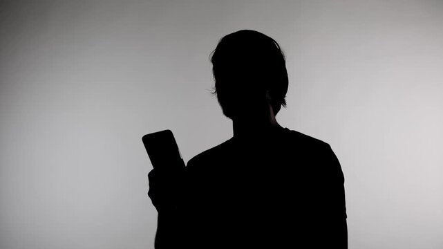 An anonymous silhouette of a man stands against a white wall and holds a phone in his hands, trying to get a network connection