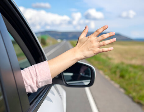 A passenger hand extends from a moving car window, playing with the wind on a sunny day along an open country road.