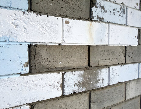 An angled close-up of a weathered masonry wall featuring alternating painted white and unpainted raw grey cement blocks with chipped and peeling paint texture.