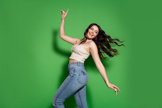 Happy young woman dancing and posing in jeans and a crop top against a bright green background, smiling with flowing hair for fashion and lifestyle portrait