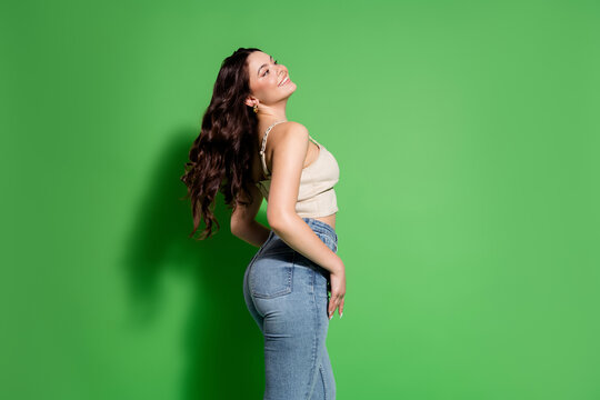 Smiling young woman in jeans and crop top posing confidently against a bright green background with flowing hair and relaxed, carefree posture