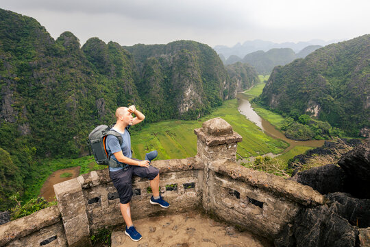 Tourist with backpack taking break to rest on rock cliff during hot day hike amidst limestone karst mountains in Ninh Binh Vietnam. 