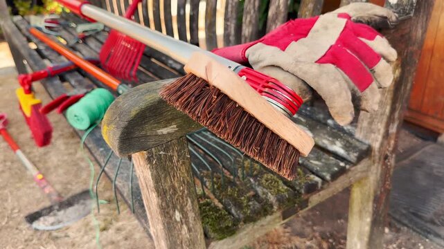 Closeup of a stiff brush head on a pole being laid down on an old wooden bench, along with other garden or yard maintenance tools and equipment.