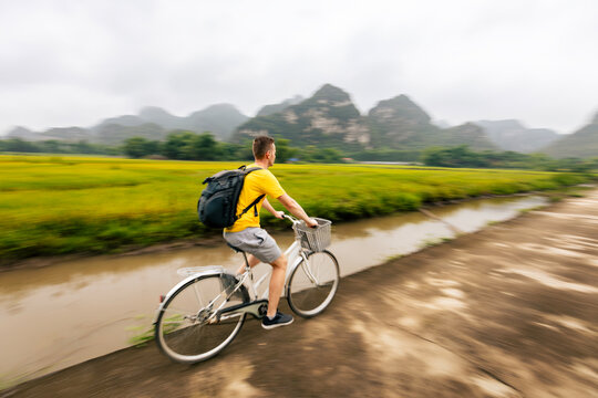 Tourist riding bicycle on road between rice fields with limestone karst mountains background in Ninh Binh Vietnam. Eco tourism, outdoor cycling adventure and rural Asian travel concept.