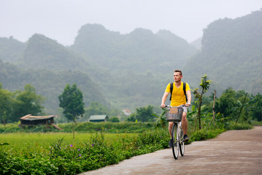 Tourist riding bicycle on road between rice fields with limestone karst mountains background in Ninh Binh Vietnam. Eco tourism, outdoor cycling adventure and rural Asian travel concept.