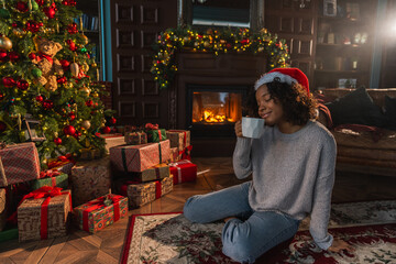 Fototapeta premium Merry Christmas. African American woman in Santa hat with cup of hot drink coffee tea near Christmas tree in dark interior. Girl in living room with Christmas tree and fireplace. Christmas eve at home
