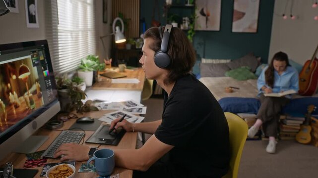 Young Asian man wearing headphones editing digital images on computer with graphics tablet while young Caucasian woman reads on bed. Perfect for remote work and freelance concepts